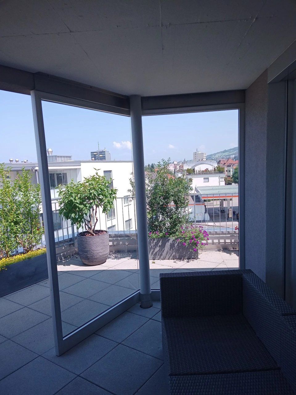 Innenansicht durch bodentiefe Fenster auf eine Terrasse, Bäume und Gebäude draussen.