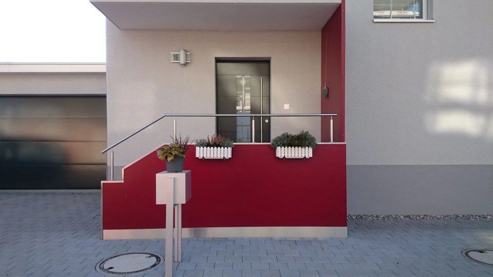 Gebäudeeingang mit roter Rampe, Metallgeländer und Topfblumen vor einer grauen Fassade.