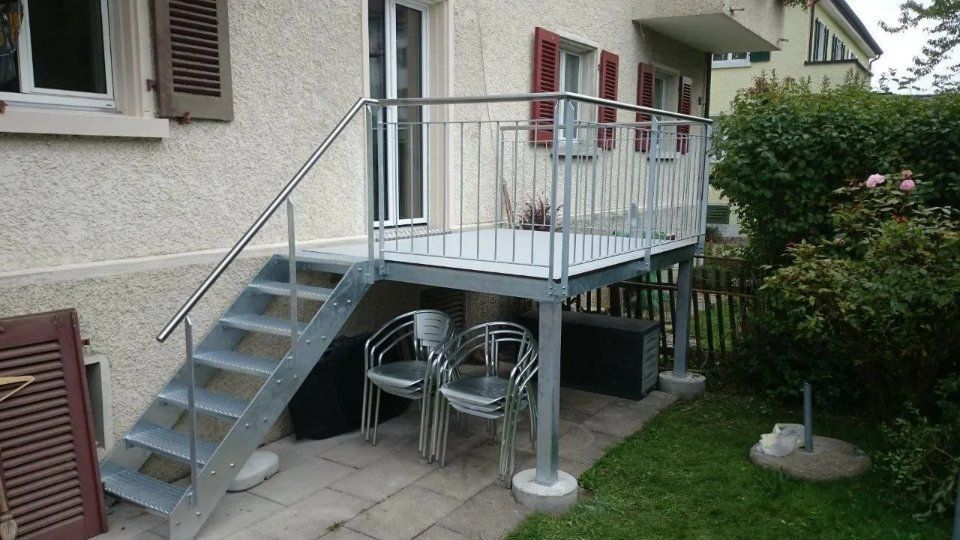 Kleine Aussentreppe aus Metall mit Podest, Tisch und Stühlen neben einem Haus, auf einer Terrasse neben einem Rasen.