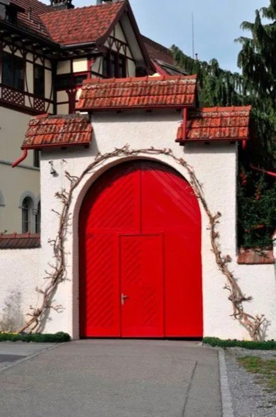 Ein leuchtend rotes Tor mit Bogen in einer weissen Mauer, dahinter ein Haus mit Ziegeldach.