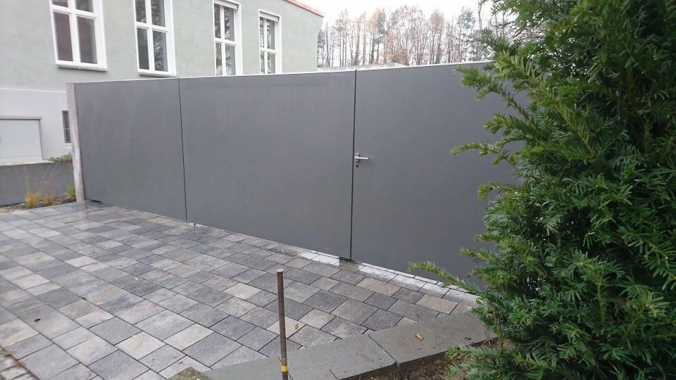 Graues Einfahrtstor neben einem gepflasterten Hof und einem Gebäude, rechts davon ein Baum.