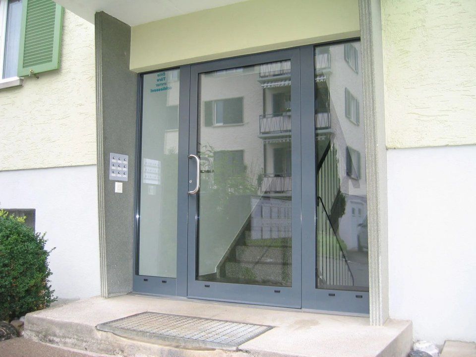 Gebäudeeingang mit gläsernen Doppeltüren, grauem Rahmen und einer kleinen Rampe mit Fussmatte am Eingang