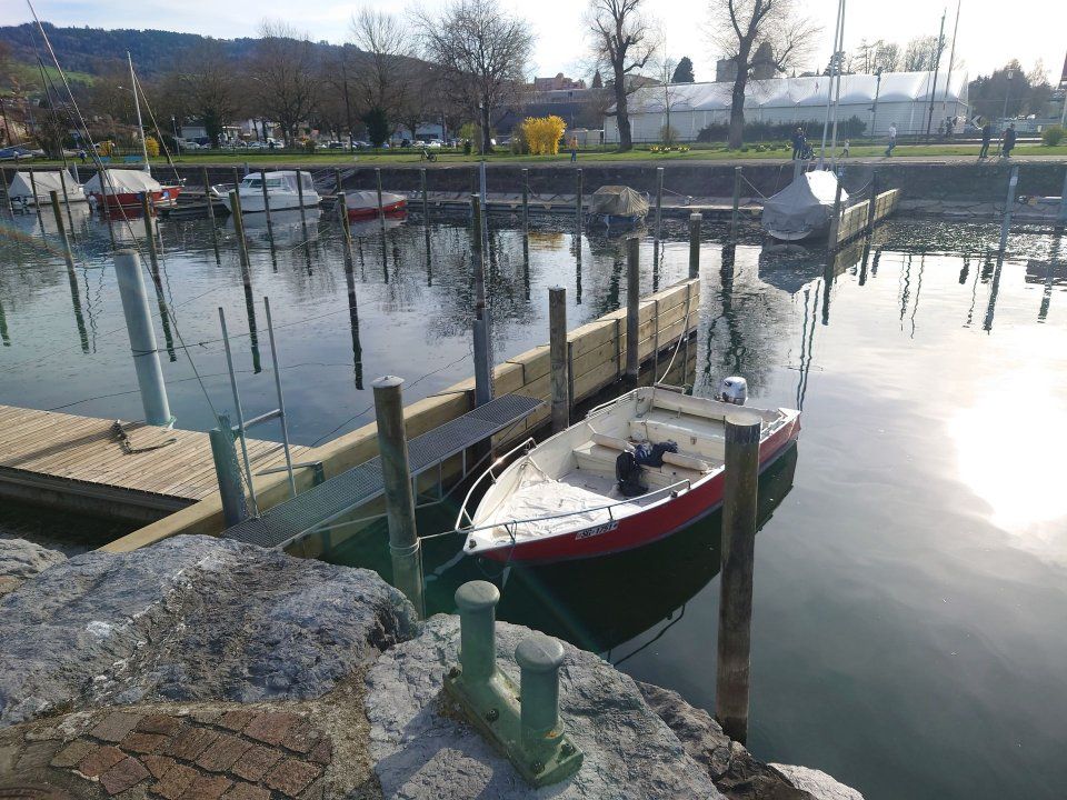 Kleines rot-weisses Boot liegt in einem ruhigen Yachthafen mit Holzstegen und hellen Wasserspiegelungen vor Anker.