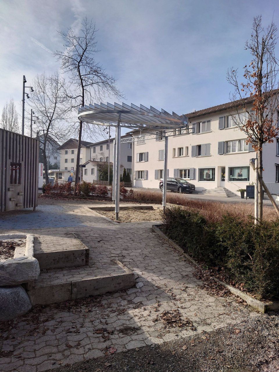 Ein Aussenhof mit Wohnhäusern, kahlen Bäumen und einer Metallpergola über einer gepflasterten Fläche.