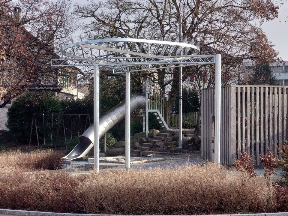 Spielplatz mit Rutsche und Klettergerüst im Freien, mit Metalldach, umgeben von Bäumen und einem Zaun.