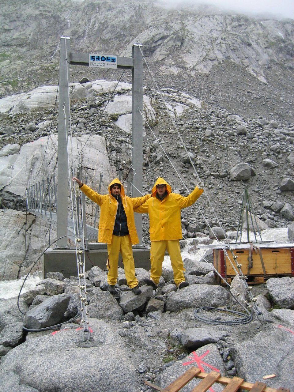 Zwei Arbeiter in gelber Regenkleidung stehen mit erhobenen Armen auf Felsen an einem schneebedeckten Bergsteigergebiet.