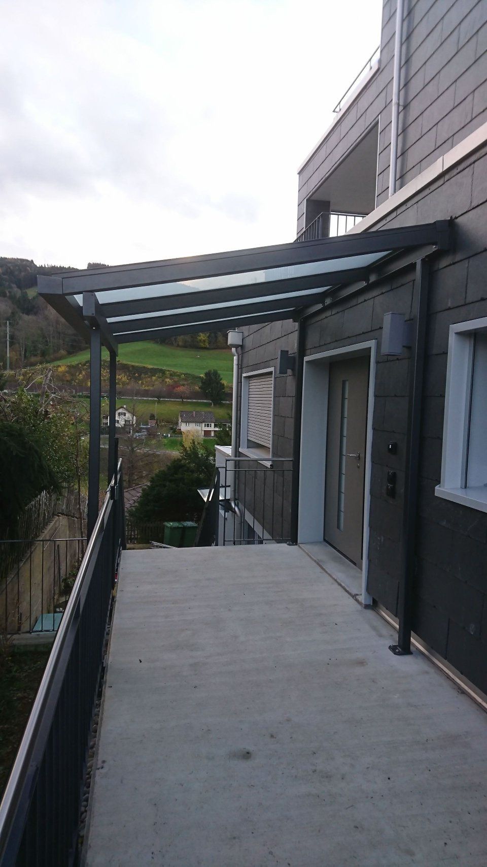 Überdachter Aussenweg neben einem Haus mit Betonweg und Metallgeländer mit Blick auf einen Hügel.