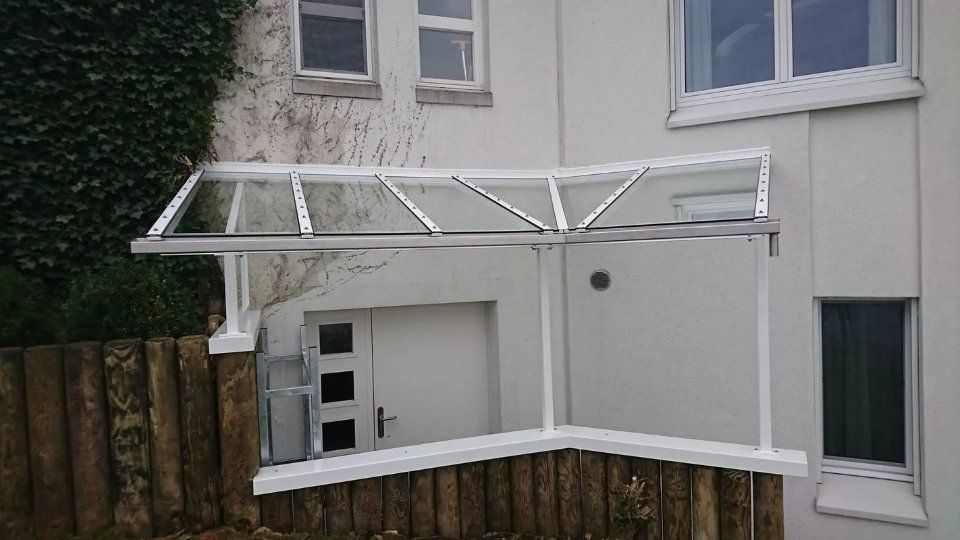 Ein mit einer weissen Markise überdachter Balkon oder eine Veranda, die an ein beigefarbenes Haus über einem Holzzaun angebaut ist.