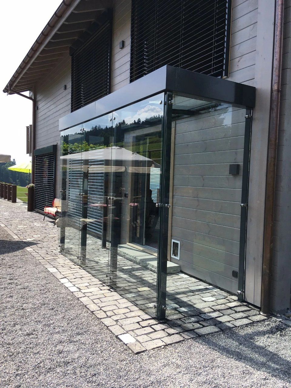 Eingang mit Glasvordach an einem grauen Backsteingebäude mit Kopfsteinpflasterweg