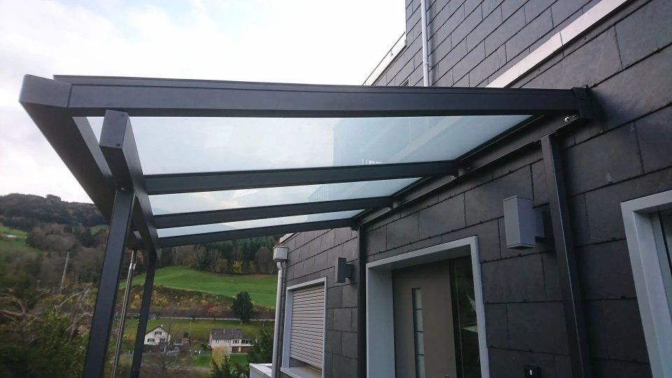 Moderne Glasmarkise über einem grauen Haus, im Hintergrund ein grüner Hügel.