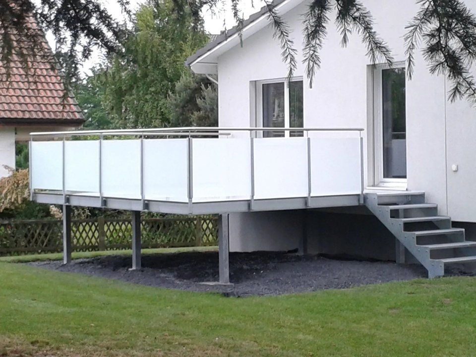 Erhöhte weisse Terrasse mit Geländer und Treppe, die an ein Haus in einem grasbewachsenen Garten angebaut ist