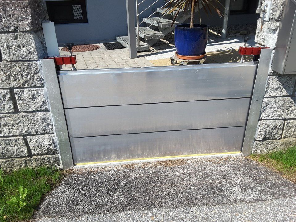 Ein graues Metalltor mit einem blauen Pflanzkübel darauf, eingefasst zwischen Steinmauern am Hauseingang.