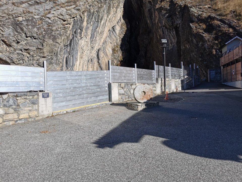 Felsiger Strassenrand mit Metallzaun und einer kreisförmigen Betonbarriere neben einem Tunneleingang