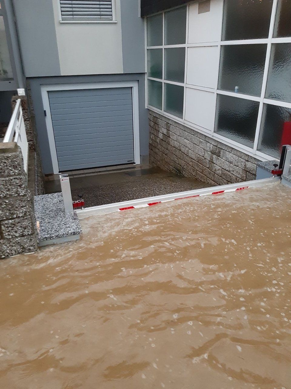 Überfluteter Innenhof neben einem Gebäude, wobei schlammiges Wasser über eine rot-weisse Absperrung steigt.