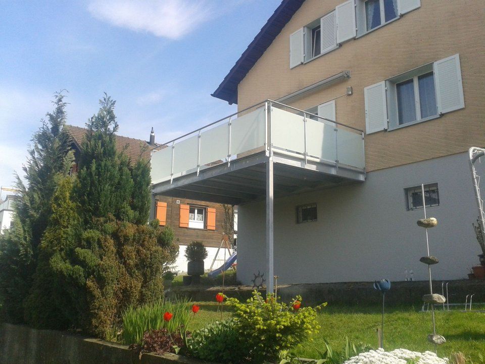 Vorstadthaus mit weissem Balkon über einem Garten und Sträuchern an einem sonnigen Tag