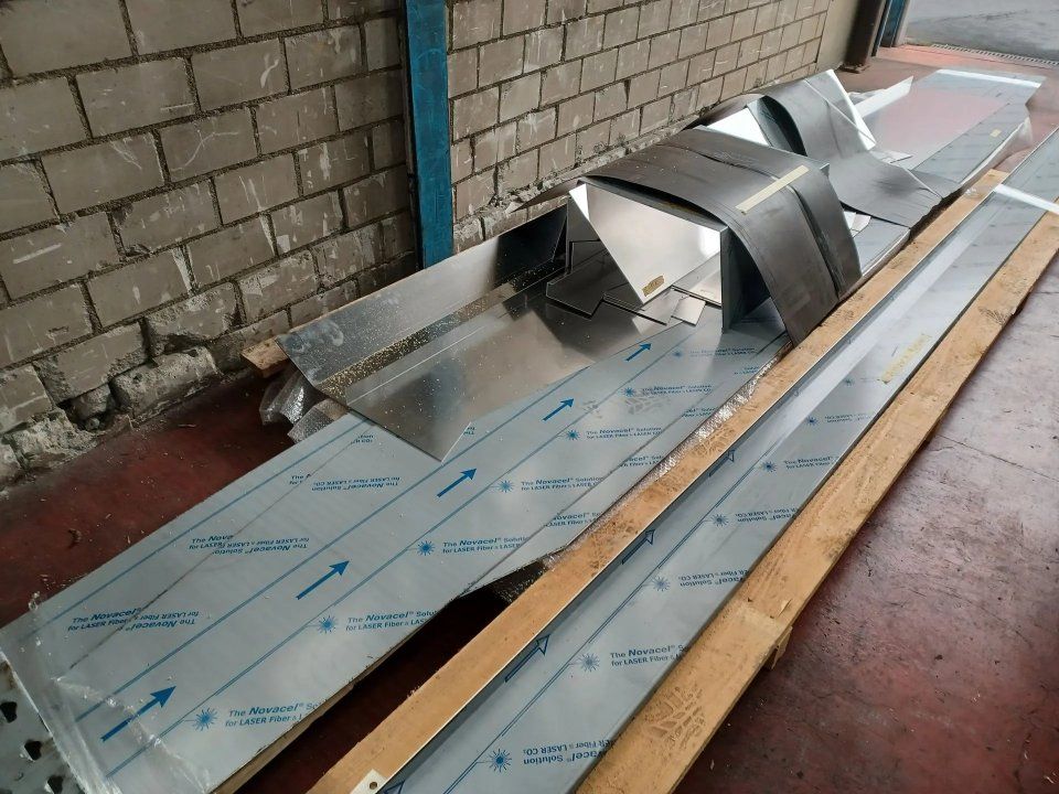 Metallene Lüftungskanalabschnitte liegen in einer Werkstatthalle auf Schutzfolien, daneben Holzbretter.