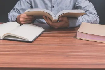 Persona leyendo un libro abierto en un escritorio de madera, con otros dos libros cerca.