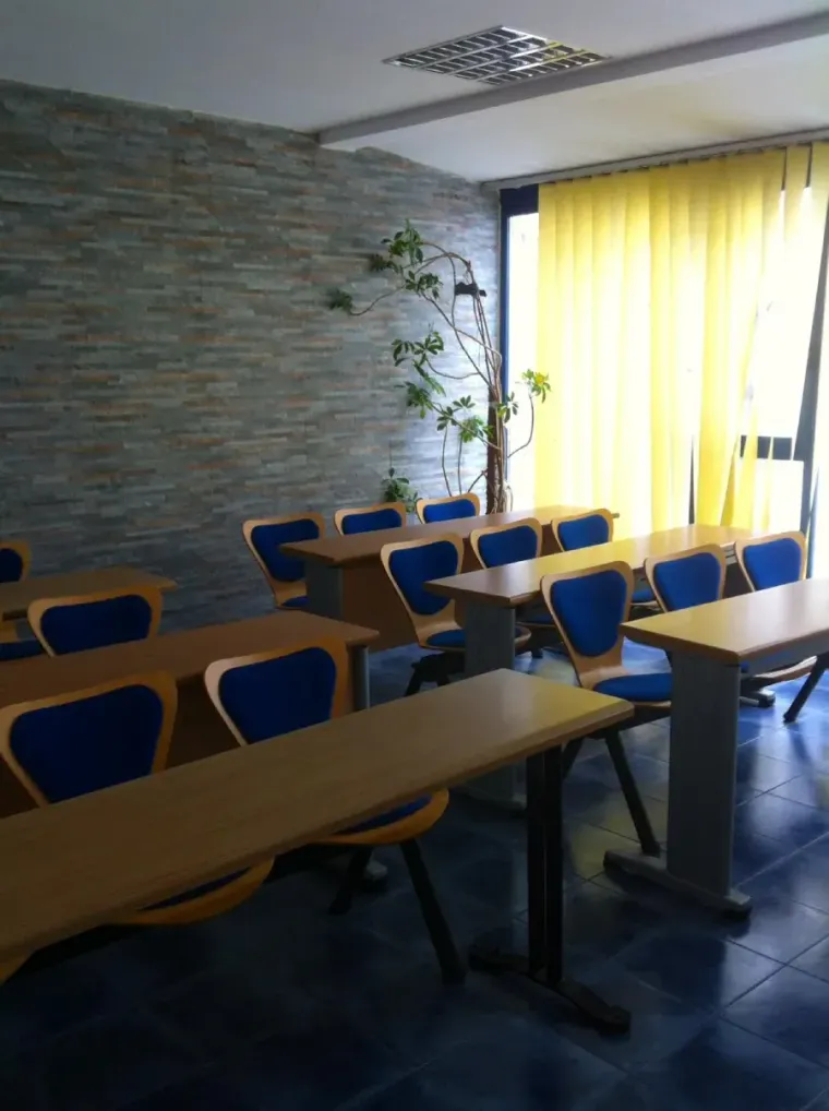 Aula con sillas azules, pupitres de madera y pared de piedra. Cerca de una ventana hay cortinas amarillas y una planta.