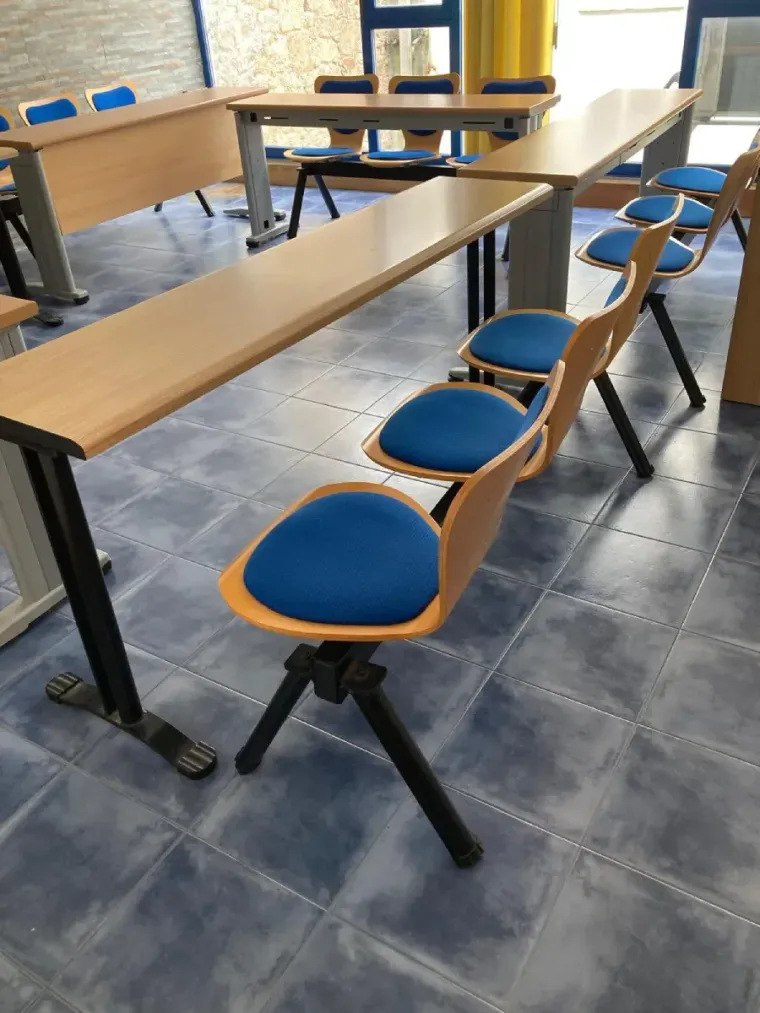 Aula con sillas azules, pupitres de madera y pared de piedra. Cerca de una ventana hay cortinas amarillas y una planta.