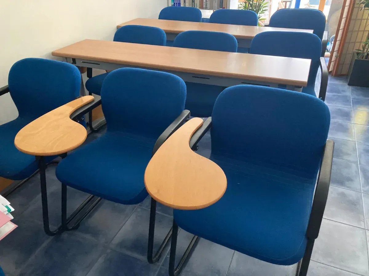 Sillas azules con reposabrazos incorporados en un aula, dispuestas cerca de una mesa larga.