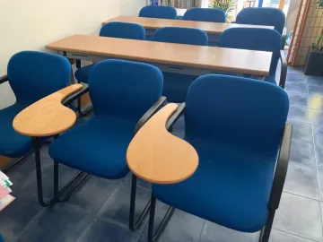Sillas azules con reposabrazos incorporados en un aula, dispuestas cerca de una mesa larga.