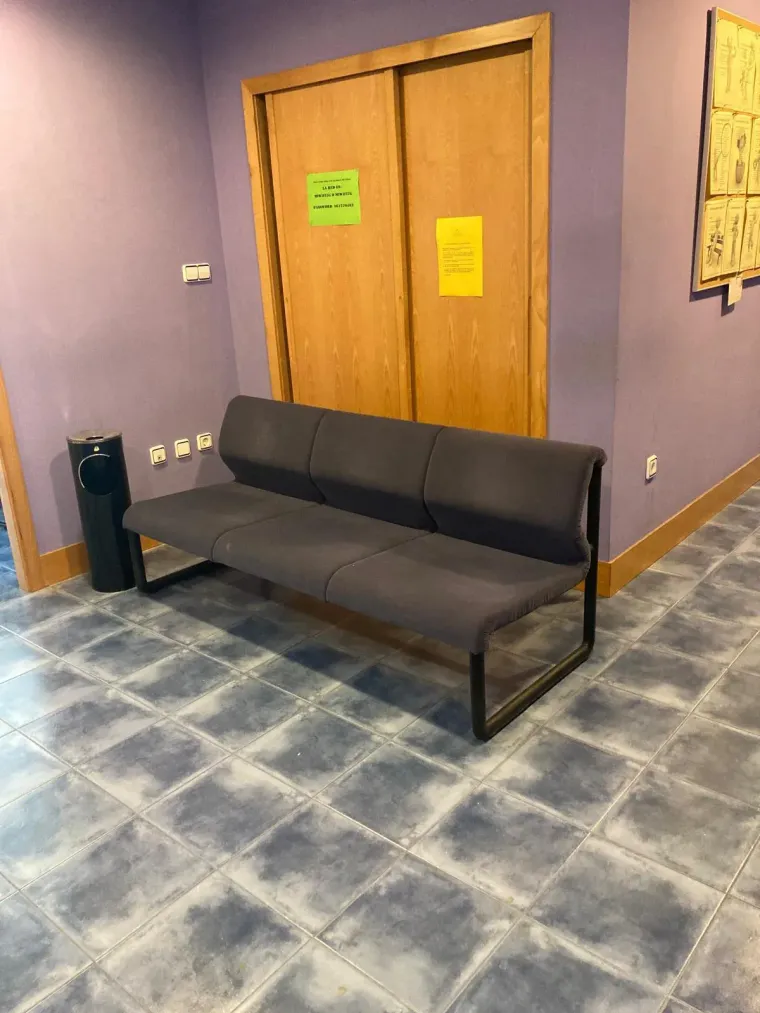 Aula con sillas azules, pupitres de madera y pared de piedra. Cerca de una ventana hay cortinas amarillas y una planta.