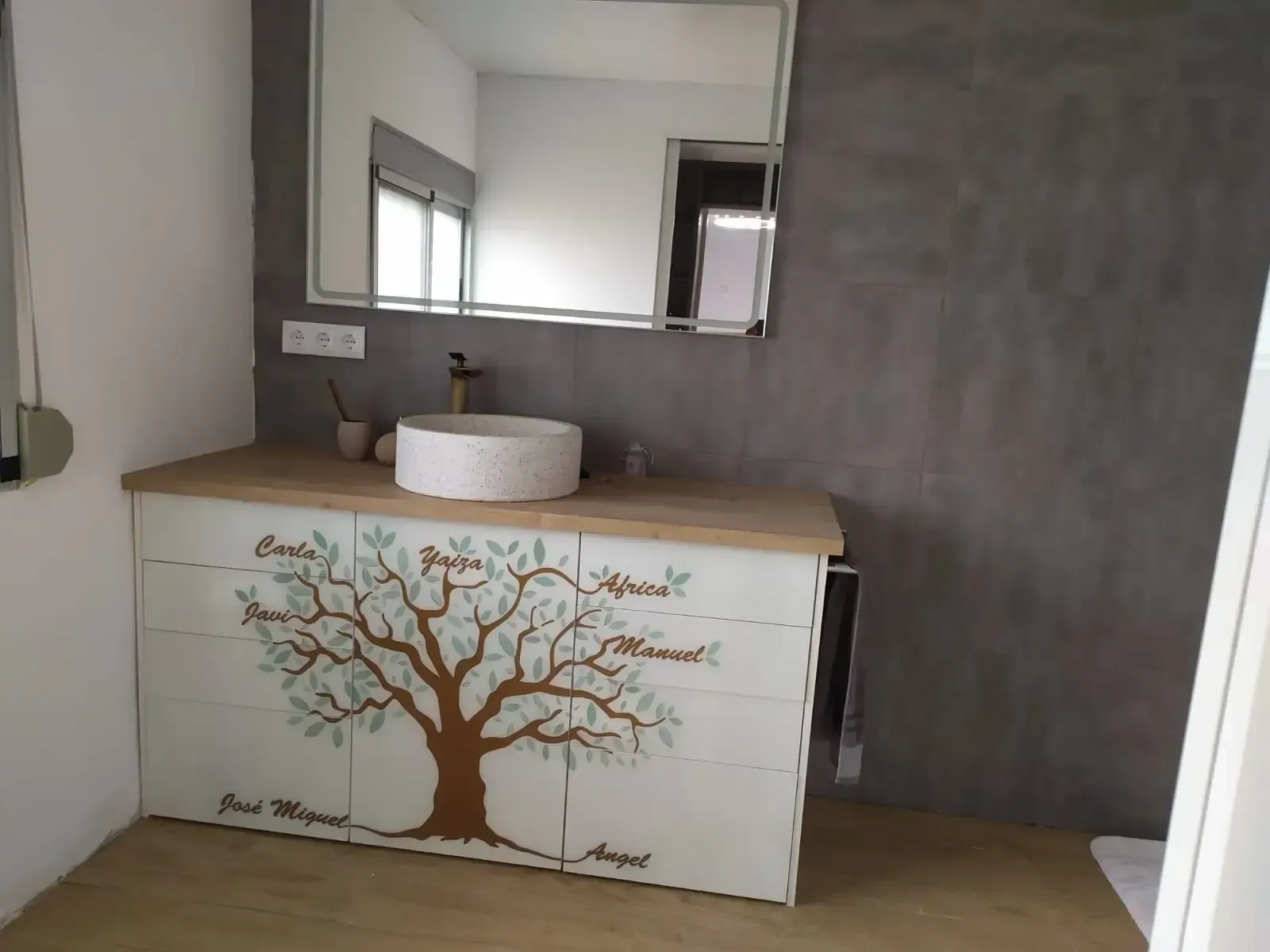 Un baño con lavabo, espejo y tocador con un árbol pintado.