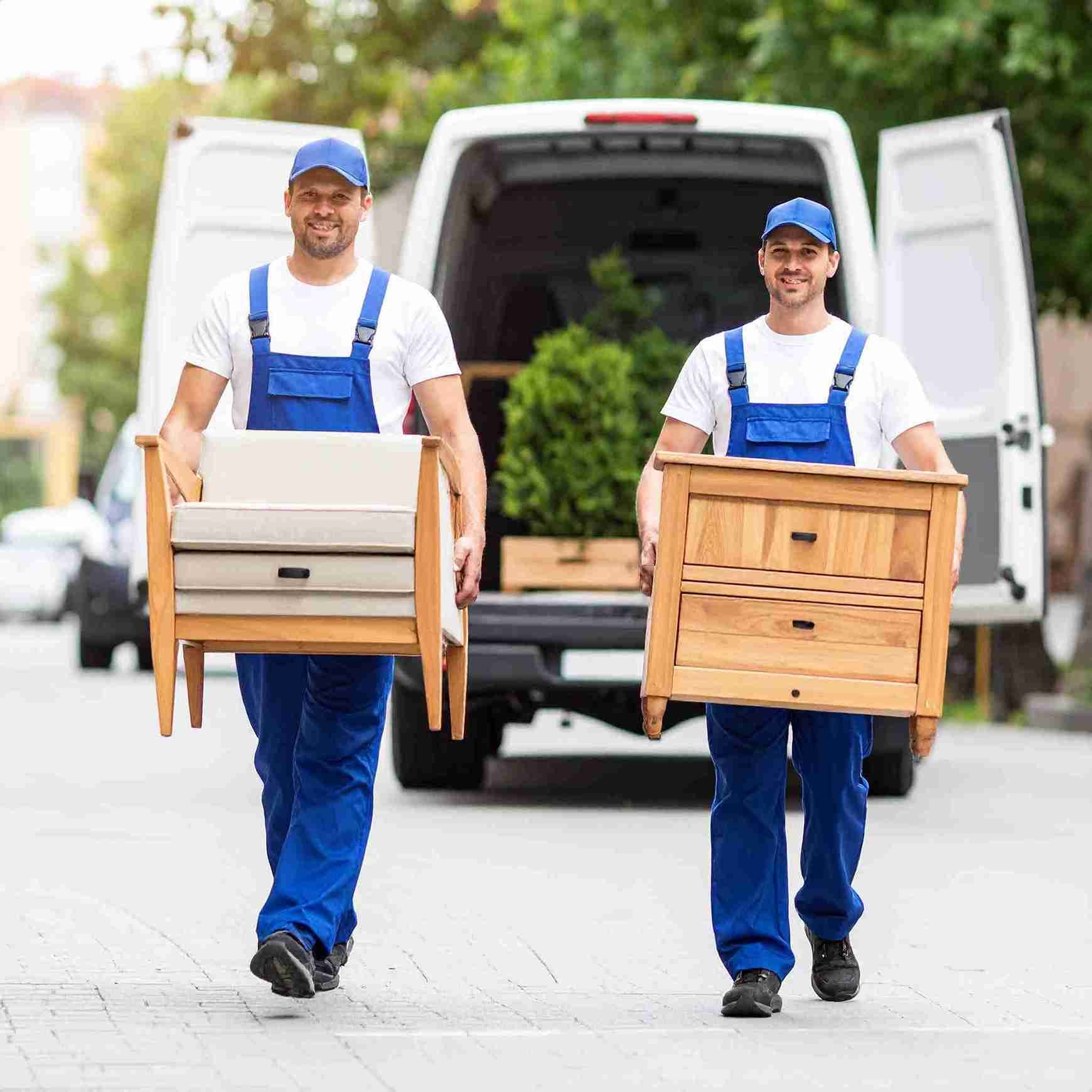 Dos trabajadores de mudanzas con monos y gorras azules transportan muebles.