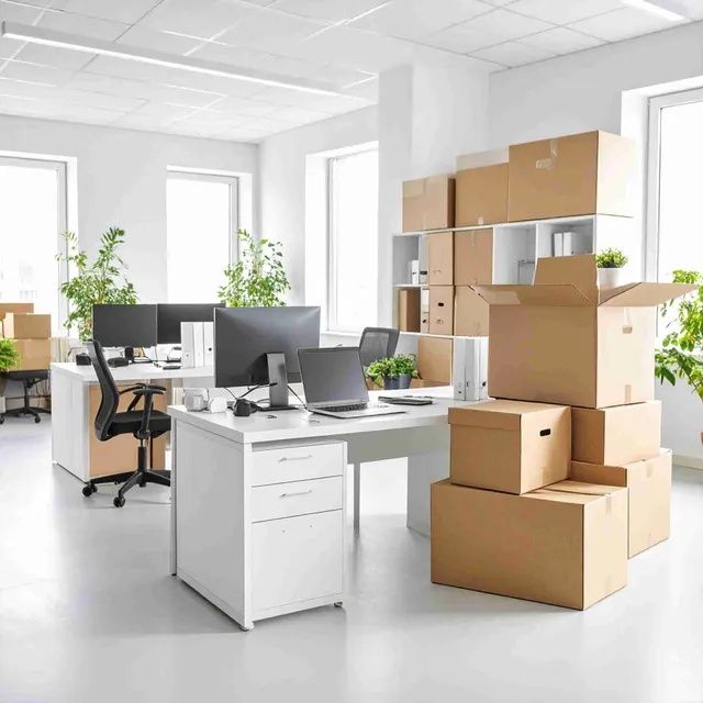 Un espacio de oficina con escritorios y monitores de computadora, lleno de cajas de cartón, lo que sugiere una mudanza.