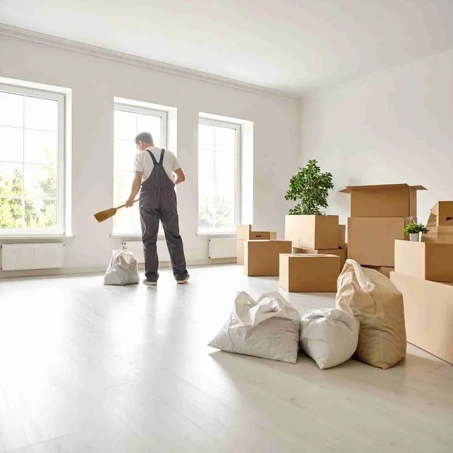 Una persona con overol barre una habitación vacía y luminosa con cajas y bolsas.