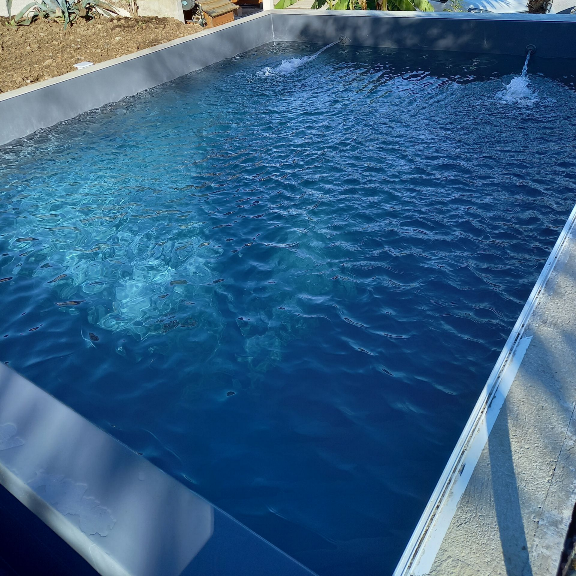 Bassin rectangulaire rempli d'eau bleue ; deux fontaines créent des bulles. Bordure en béton. Aménagement extérieur.