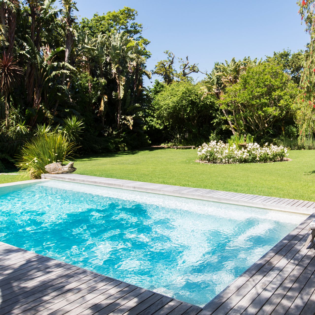 Piscine rectangulaire aux eaux turquoise, entourée d'une terrasse en bois et d'une pelouse verdoyante, avec un jardin luxuriant en arrière-plan.