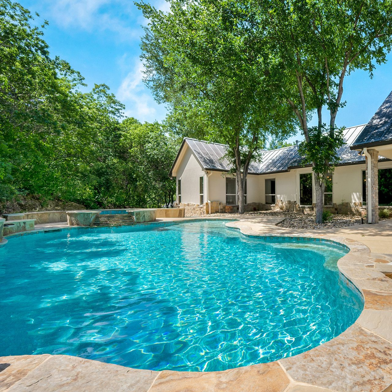 Piscine dans un jardin, avec une maison en arrière-plan et des arbres tout autour, sous un ciel lumineux.