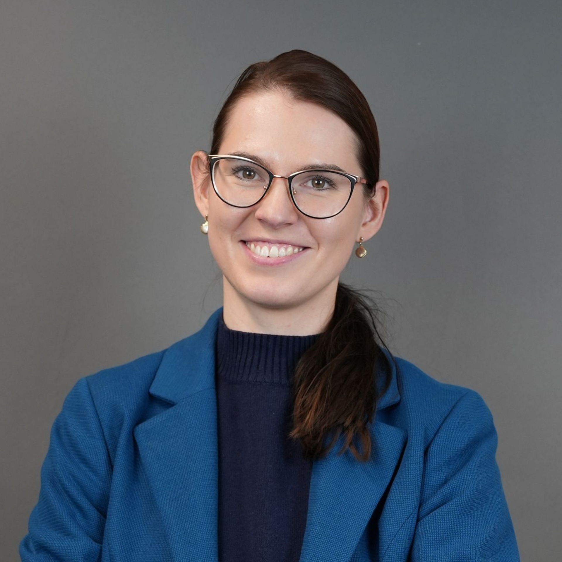 Woman with glasses, smiling, wearing a blue blazer and sweater, posed against a gray backdrop.