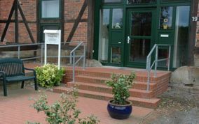 Außenansicht eines Gebäudes mit Backsteintreppe, grüner Tür und Fachwerk. Es gibt eine Rollstuhlrampe, eine Bank und Topfpflanzen.