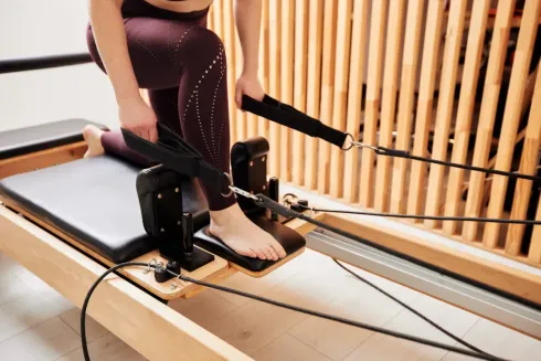 Persona haciendo Pilates en una máquina reformer, tirando de correas, con una pared de madera de fondo.