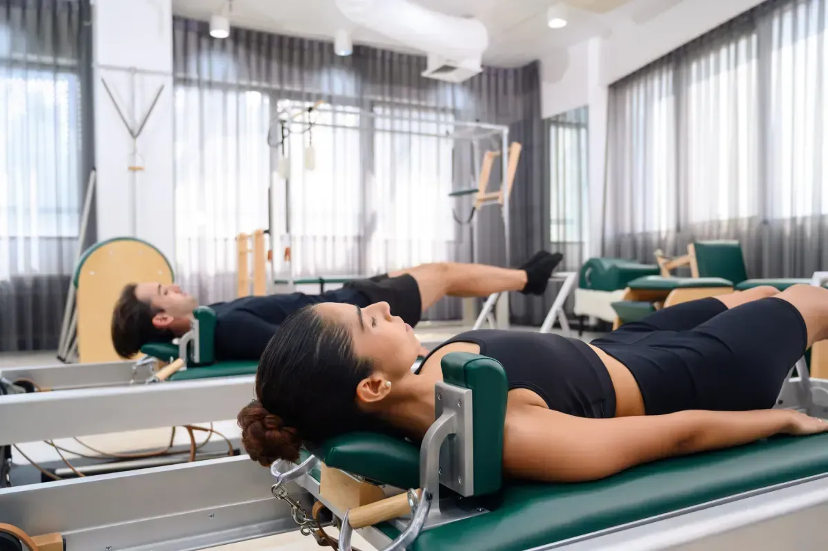 Dos personas en máquinas de Pilates Reformer. Están tumbadas boca arriba con las piernas levantadas, dentro de un estudio.