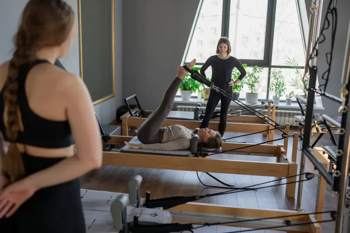Una mujer practica Pilates en un aparato, mientras el instructor la observa. Otra mujer observa en un estudio iluminado.
