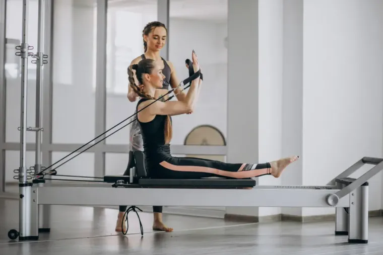 Mujer haciendo Pilates con instructor en máquina reformer.