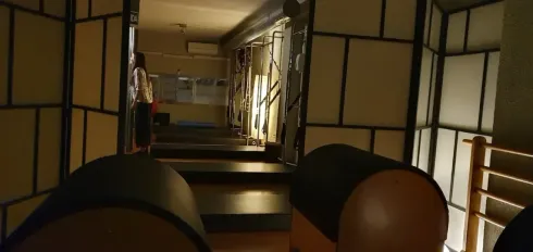 Un interior tenuemente iluminado con pasillo, escaleras y tabiques. Se ven dos objetos redondos en primer plano.