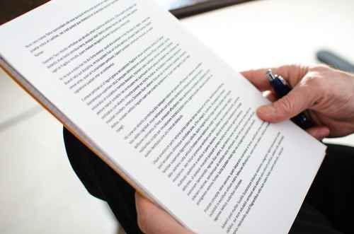 Pessoa segurando e lendo um documento; caneta na mão; papel branco com texto preto.