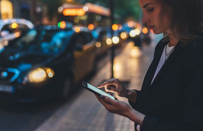 Une femme utilise son téléphone dans une rue de la ville, la nuit, avec des voitures et des lumières en arrière-plan.