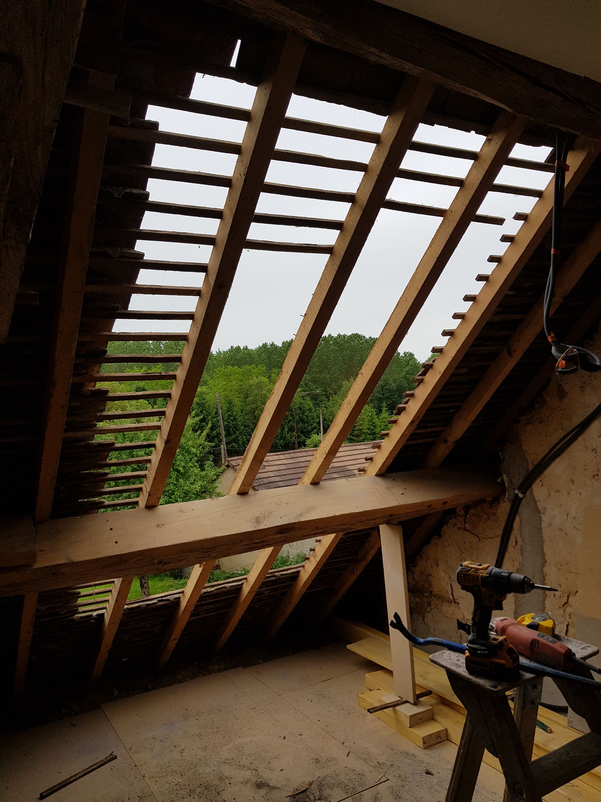 Vue depuis un grenier partiellement aménagé avec poutres apparentes, ciel et arbres. Des outils sont visibles sur un établi.