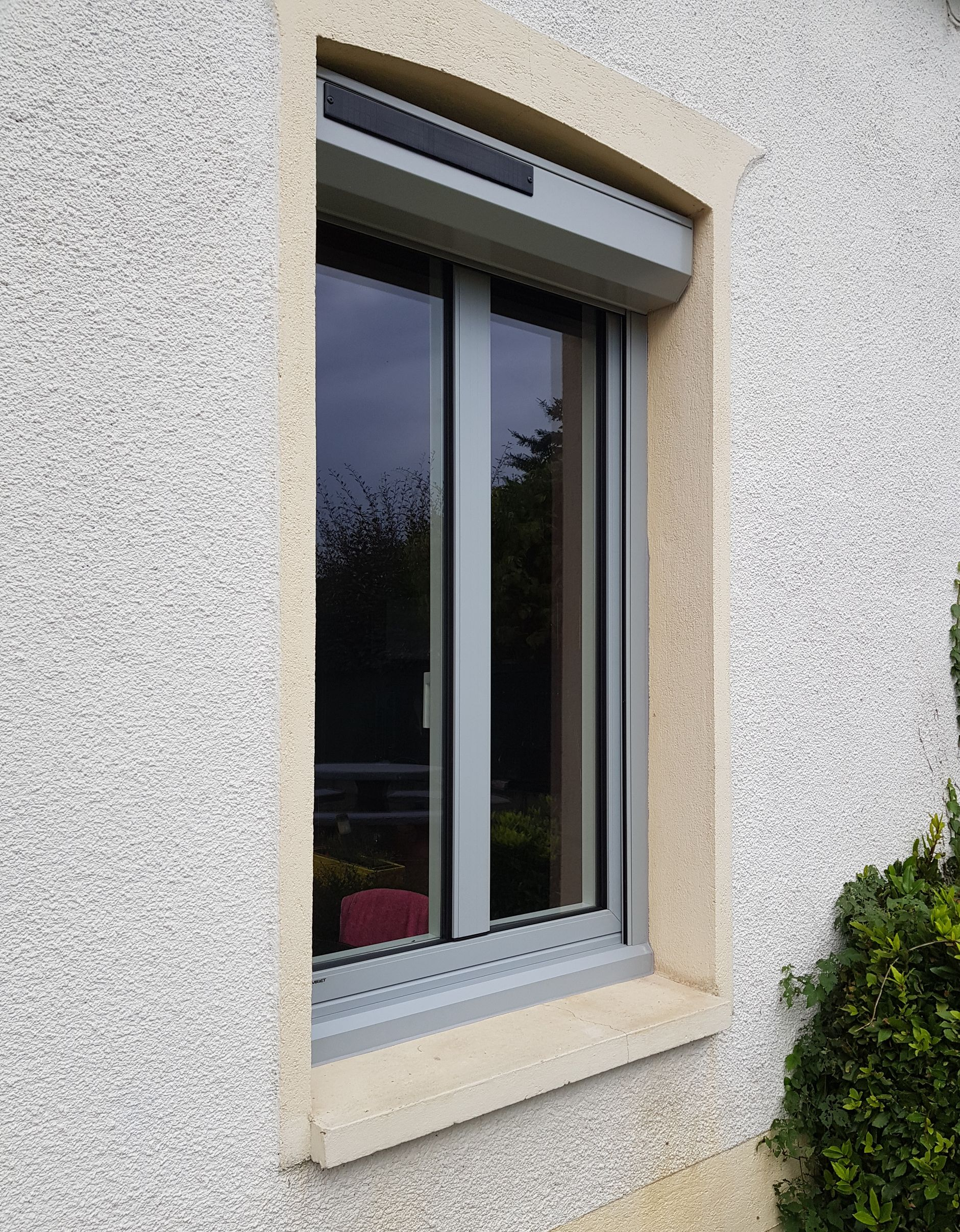 Fenêtre avec volet roulant gris fermé, dans un encadrement en pierre claire, encastré dans un mur en stuc.