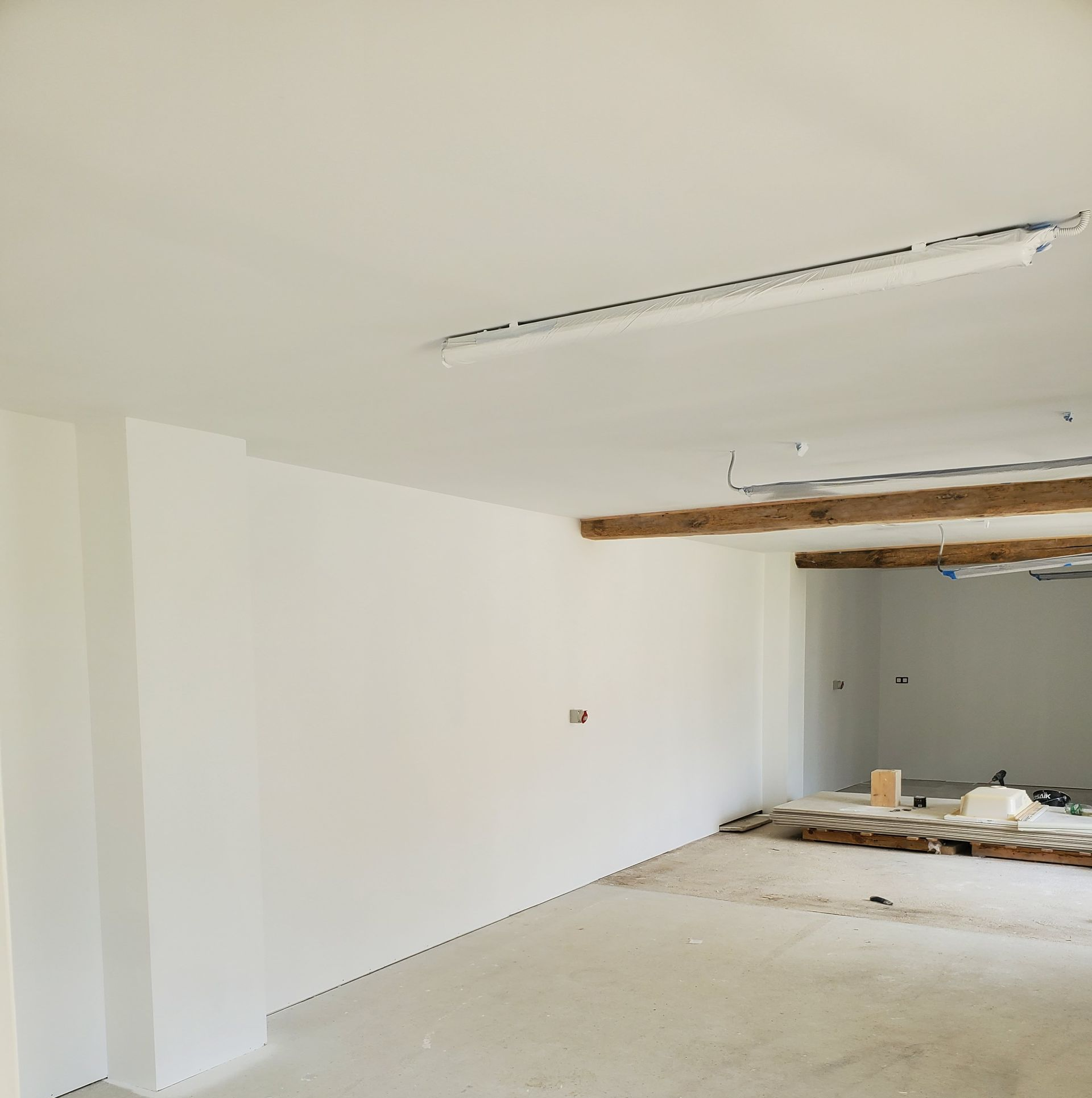 Vue intérieure d'une pièce en rénovation, avec murs blancs, poutres apparentes et sol en béton.