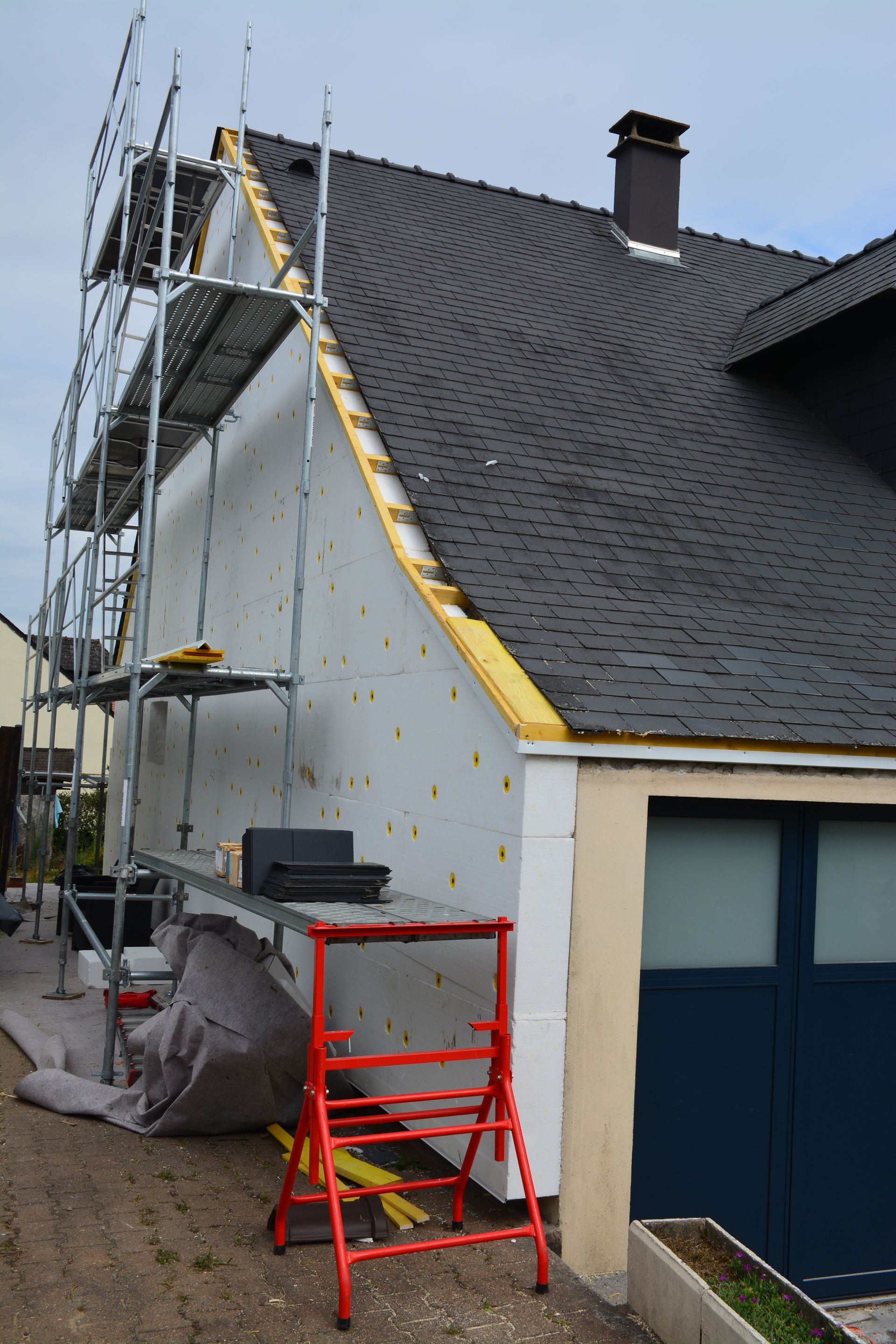 Maison en construction avec échafaudage. Mur blanc isolé, toit foncé et garage.