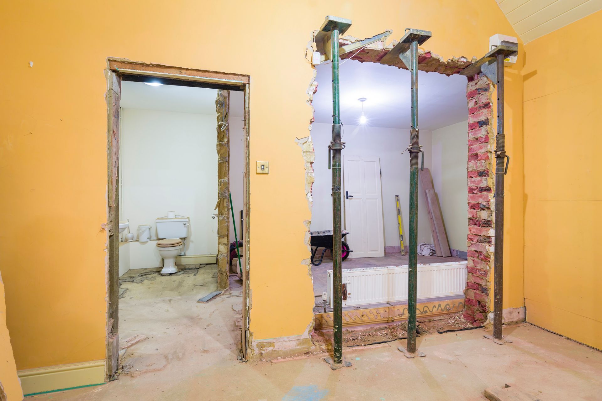 Chambre en rénovation : porte d'entrée vers une salle de bains à gauche, porte ouverte avec supports à droite. Murs jaunes, débris de construction.
