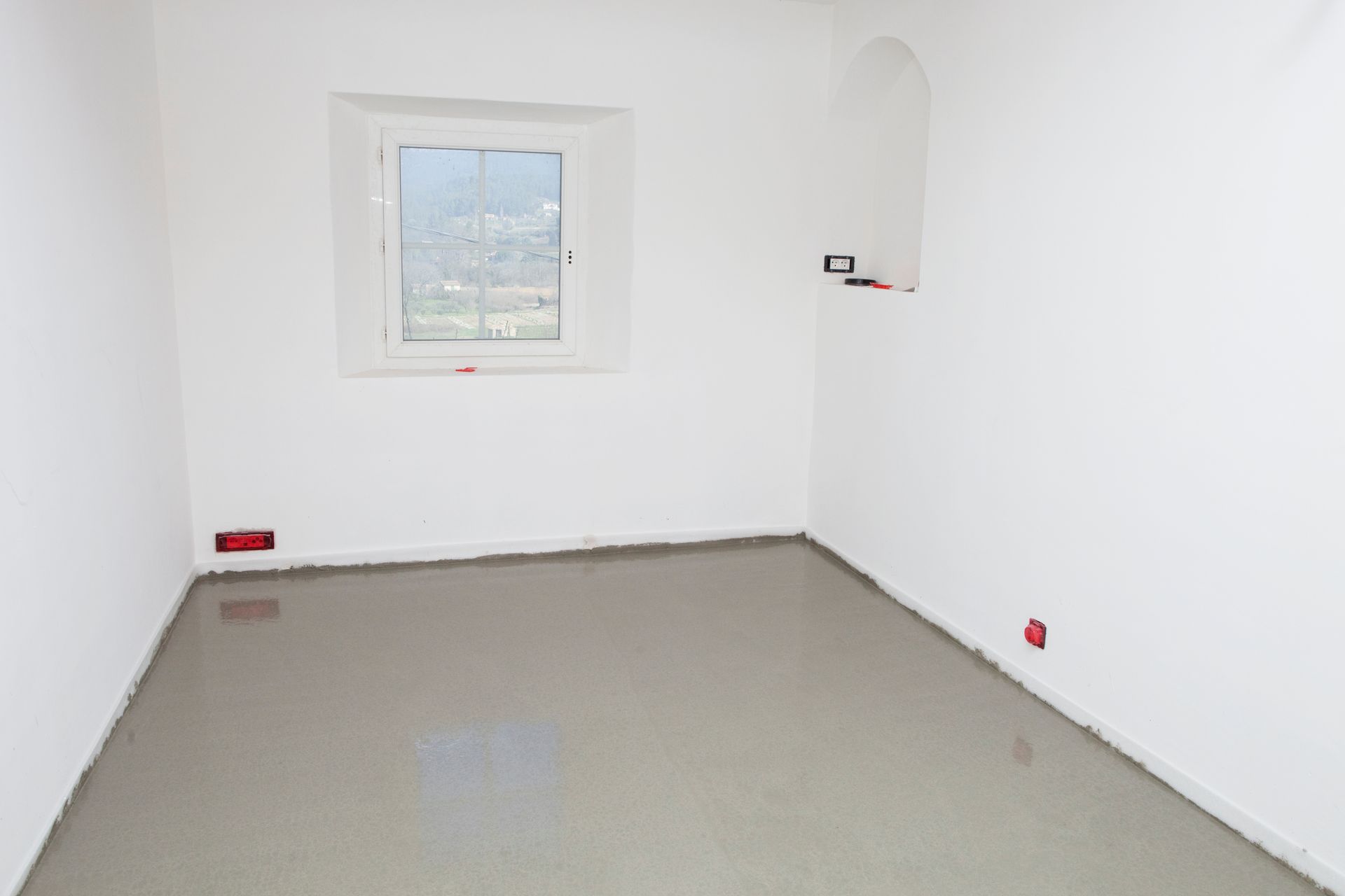 Pièce vide avec sol en béton gris, murs blancs, petite fenêtre et prises électriques.