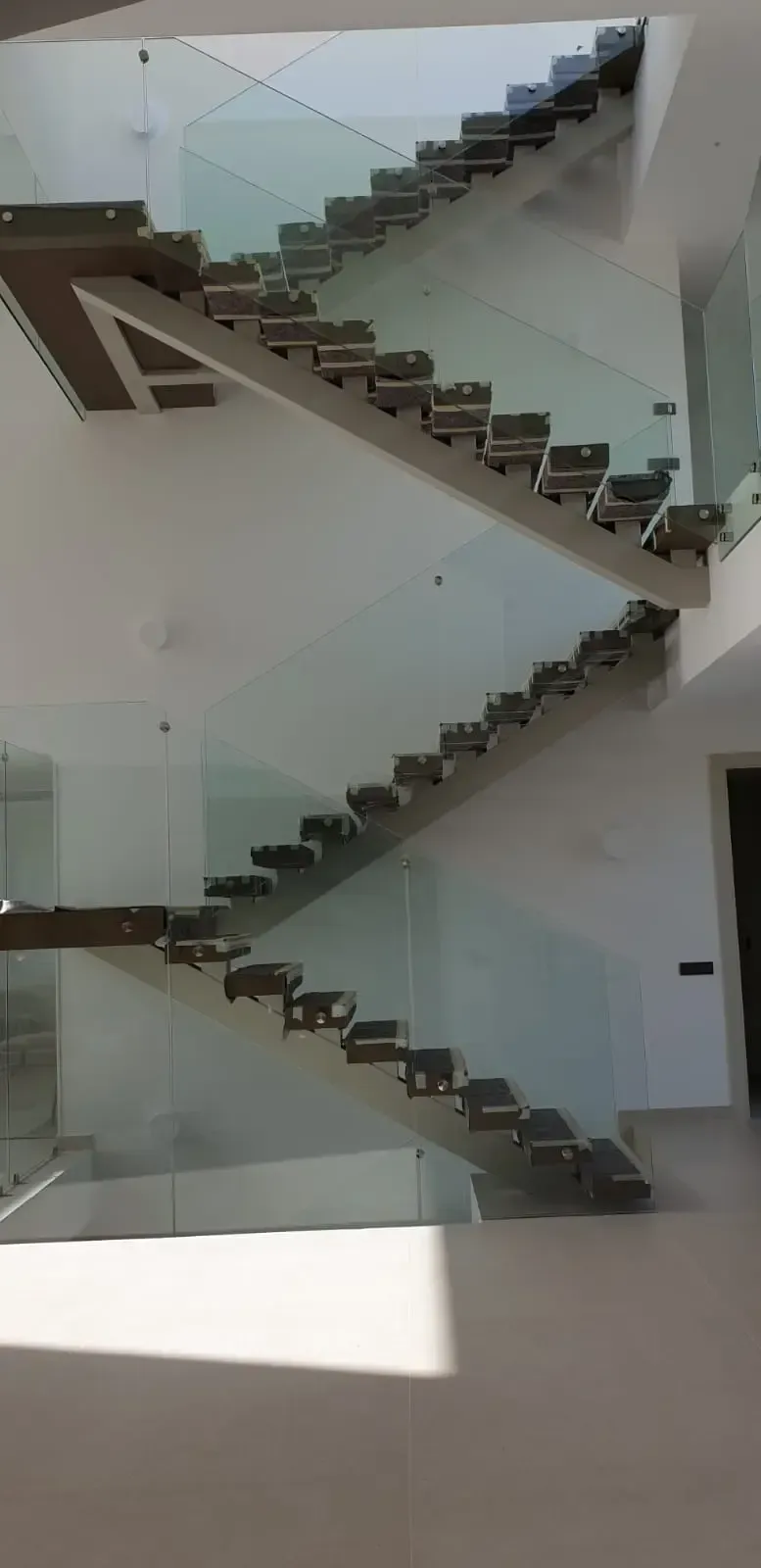 Escalera moderna con peldaños de madera oscura, estructura de largueros a la vista y barandilla de cristal sin marco en una habitación blanca.