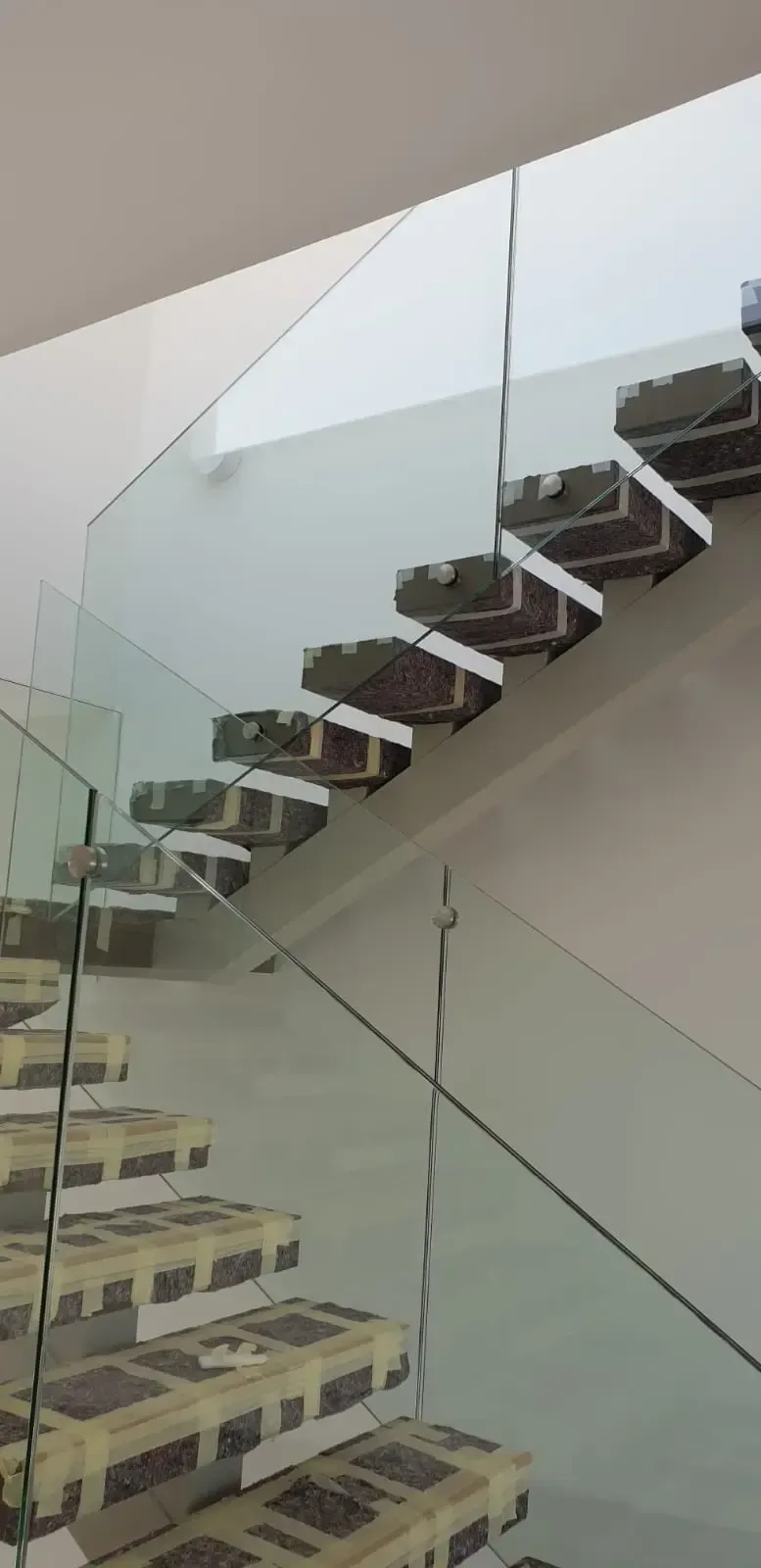 Una escalera moderna en construcción con cinta protectora en los peldaños oscuros y barandillas de cristal fijadas a un lado.